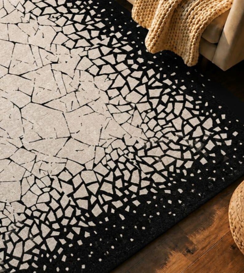 May include: A rectangular rug with a unique design. The rug features a pattern of black geometric shapes against a beige background. The design transitions from a dense black border to a lighter, fragmented pattern in the center. A tan blanket is draped over a sofa in the background.