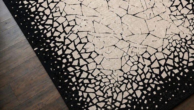May include: A black and beige area rug with a mosaic pattern. The rug's design features a gradient effect, transitioning from a solid black border to a beige center with fragmented shapes. The rug is placed on a dark wood floor.