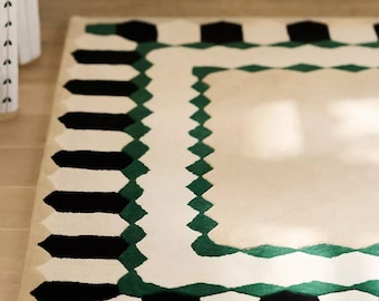 May include: A rectangular area rug featuring a cream-coloured centre bordered by a geometric pattern of alternating black and dark green shapes. The outer edge displays a repeating sequence of black and cream vertical bars with green accents.