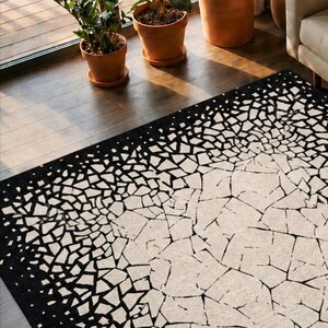 May include: A rectangular area rug with a black border and a white and black mosaic pattern. The rug's design transitions from a solid black border to a fragmented, geometric pattern in the center. The rug is on a wooden floor.