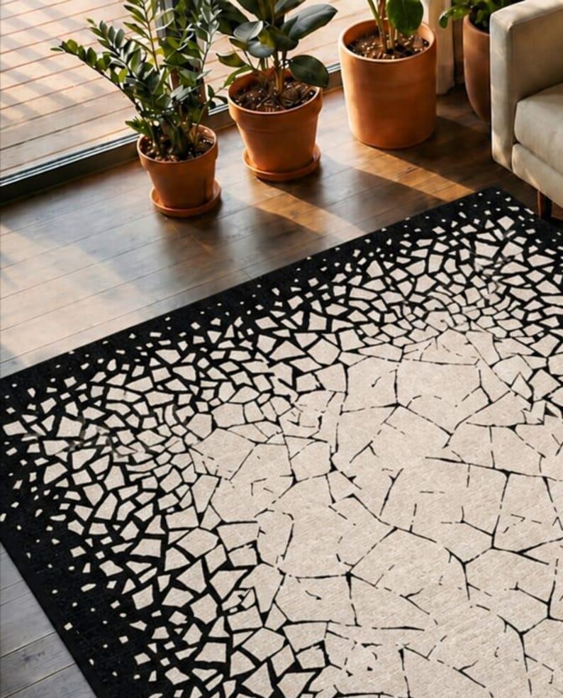 May include: A rectangular area rug with a black border and a white and black mosaic pattern. The rug's design transitions from a solid black border to a fragmented, geometric pattern in the center. The rug is on a wooden floor.