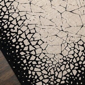 May include: A black and beige area rug with a mosaic pattern. The rug's design features a gradient effect, transitioning from a solid black border to a beige center with fragmented shapes. The rug is placed on a dark wood floor.