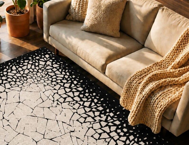 May include: A beige sofa with two throw pillows and a chunky knit blanket. A black and white geometric patterned rug is in the foreground. Potted plants are visible in the background on a wooden floor.