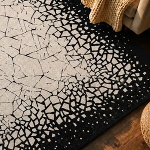 May include: A rectangular rug with a unique design. The rug features a pattern of black geometric shapes against a beige background. The design transitions from a dense black border to a lighter, fragmented pattern in the center. A tan blanket is draped over a sofa in the background.