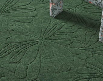 May include: A deep forest green area rug featuring a large-scale, tone-on-tone embossed floral pattern with carved petal details. The rug has a thick, plush texture and is partially covered by the legs of a chair with a light blue bird print.