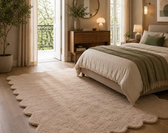 May include: A large, rectangular area rug with a scalloped edge design. The rug features a solid, light beige colour and a thick, plush, textured pile surface. It is positioned on a light-toned wooden floor, extending from the foreground.