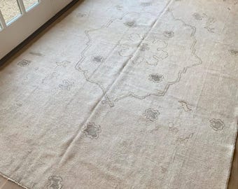 May include: A large, rectangular rug with a faded, vintage appearance. The rug is primarily beige with a central medallion and scattered floral motifs in a muted grey-brown. The rug is placed on a wooden floor.