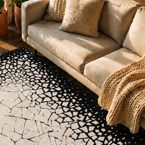 May include: A beige sofa with two throw pillows and a chunky knit blanket. A black and white geometric patterned rug is in the foreground. Potted plants are visible in the background on a wooden floor.