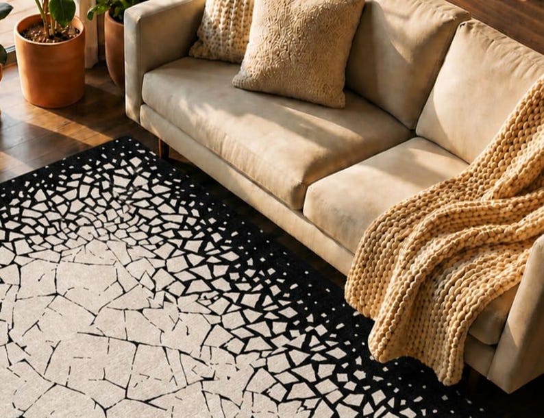 May include: A beige sofa with two throw pillows and a chunky knit blanket. A black and white geometric patterned rug is in the foreground. Potted plants are visible in the background on a wooden floor.