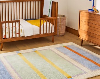 May include: A rectangular area rug featuring a geometric pattern of light green, blue, and cream sections accented by bold yellow and orange vertical stripes. The rug is positioned on a light wood floor in front of a wooden crib and a wooden dresser.