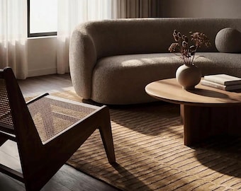 May include: A contemporary living room featuring a curved, taupe sofa, a circular wooden coffee table, and a woven chair. A beige rug with linear designs covers the floor. A vase with dried flowers sits on the table, and a window with sheer curtains.