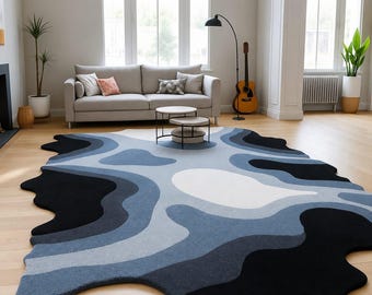 May include: An area rug with an irregular, organic shape featuring a layered, topographical pattern in shades of black, dark blue, light blue, and white. The rug has a textured, tufted surface and covers a portion of the light-coloured wooden floor.