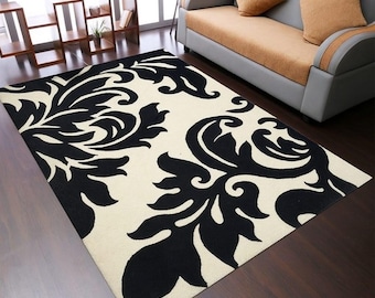 May include: A rectangular rug featuring a cream-coloured base with a bold, repeating black floral design. The rug is positioned on a dark wooden floor, enhancing the contrast. A sofa and side tables are visible in the background.