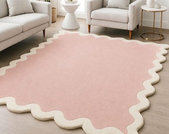 May include: A rectangular area rug featuring a solid light pink centre and a thick, cream-coloured scalloped border. The rug has a soft, plush texture and is laid flat on a light-toned wooden floor, showcasing a wavy edge design throughout.