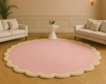 May include: A large, circular area rug featuring a solid light pink centre and a thick, scalloped cream-coloured border. The rug has a soft, textured surface and is positioned flat on a light brown wooden floor in a room with two white sofas.