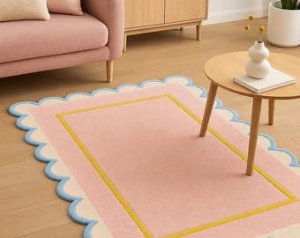 May include: A pink rectangular rug with a scalloped light blue edge. The rug features a cream border and a yellow rectangular design in the centre. It's placed on a wooden floor, with a pink sofa and wooden furniture visible in the background.