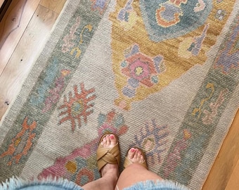 May include: A rectangular area rug featuring a central medallion and geometric border patterns in muted tones of mustard yellow, dusty blue, sage green, and soft pink on a light beige background with a textured, woven pile construction.