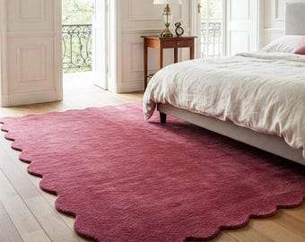 May include: A large, rectangular area rug in a deep, solid berry-red colour. The rug features a distinct scalloped edge design along its entire perimeter, creating a soft, wavy border that contrasts with the straight lines of the wooden floor.
