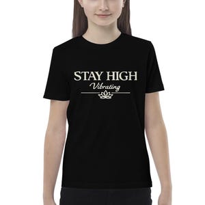 Pode incluir: T-shirt preto com o texto branco "STAY HIGH Vibrating" e um gráfico de flor de lótus. A t-shirt é de gola redonda.