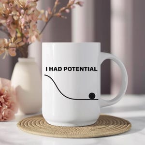 Puede incluir: Una taza de cerámica blanca con el texto negro I HAD POTENTIAL sobre un gráfico de línea negra que representa una curva descendente que termina en una sección plana con un pequeño círculo negro en la parte inferior de la curva a la derecha.