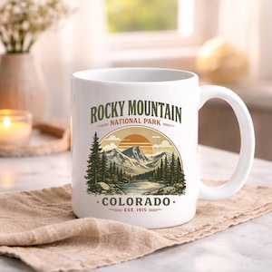 May include: A white ceramic mug featuring a graphic illustration of Rocky Mountain National Park in Colorado. The design includes a mountain range, a lake, pine trees, and a sunset, with the text Rocky Mountain National Park Colorado Est. 1915.