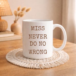 May include: A white ceramic mug with a handle, featuring the brown text "MISS NEVER DO NO WRONG" printed in a typewriter-style font on the front. The mug is cylindrical with a smooth finish and sits upright on a round, cream-colored woven coaster.