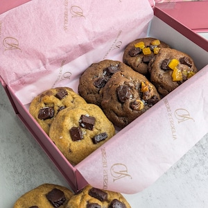 May include: A pink gift box containing several round chocolate chip cookies, some topped with orange fruit pieces. The box is lined with pink tissue paper printed with the text FOR YOUR SWEETEST MOMENTS and a logo. Two additional cookies sit outside the box.