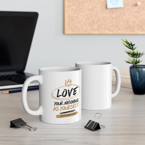 Könnte beinhalten: Zwei weiße Keramikbecher. Auf einem Becher steht in Schwarz und Gold der Text "LOVE your neighbor as yourself" mit einer Kronengrafik. Der andere Becher ist schlicht weiß. Der Text "Leviticus 19:18" ist ebenfalls auf dem Becher zu sehen.