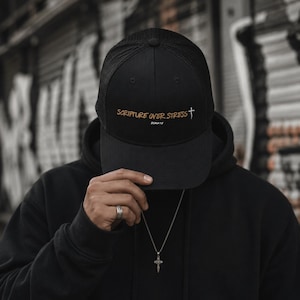 May include: Black trucker hat with the text "SCRIPTURE OVER STRESS" in gold lettering and a white cross graphic. The hat has a mesh back and a curved brim. The hat is worn by a person wearing a black hoodie.