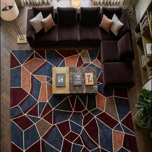 Op de afbeelding: Bovenaanzicht van een woonkamer met een bordeauxkleurige hoekbank, een geometrisch tapijt en een salontafel met marmeren blad. Het tapijt heeft blauwe, rode en oranje vormen. Neutrale kussens sieren de bank. Een vloerlamp en kroonluchter zorgen voor verlichting.