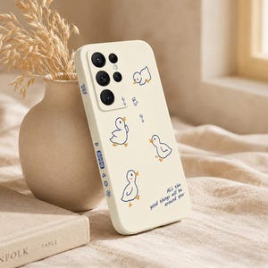 Peut inclure: Une coque de téléphone crème avec un motif ludique de canards de dessin animé et de notes de musique. La coque porte le texte "ALL the good things will be around you". La coque est à côté d'un vase avec des plantes séchées.