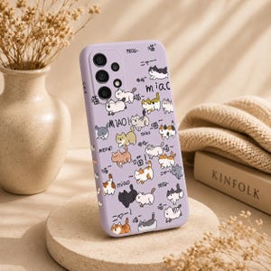 May include: A lilac phone case featuring a playful pattern of cartoon cats in various poses. The design includes Japanese text and the word "MEOW!" in English. The case is shown on a beige surface.