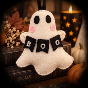 May include: A cream-colored felt ghost ornament featuring two black felt eyes, two small pink felt circles for cheeks, and a black felt banner draped across the front. The banner displays the word BOO in sparkling silver-toned letters.