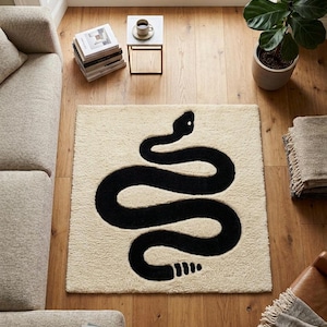 Puede incluir: Una alfombra rectangular con fondo color crema y un gran gráfico de serpiente negra centrado. La serpiente se muestra en una postura sinuosa en forma de S con un cascabel distintivo al final de la cola, confeccionada en pelo tufting.