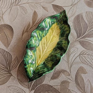 Può includere: Un piatto in ceramica a forma di foglia con finitura lucida. Il piatto presenta una vibrante tavolozza di colori, con una sezione centrale gialla e un bordo verde scuro, accentuato da macchie verdi più scure. I bordi della foglia sono delicatamente smerlati.