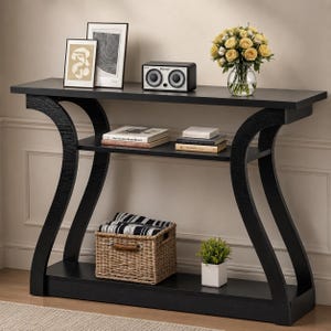 May include: Black console table with a curved design. The table has two shelves and a lower base. Decorative items include framed art, a small speaker, books, a basket, and a vase of yellow flowers.
