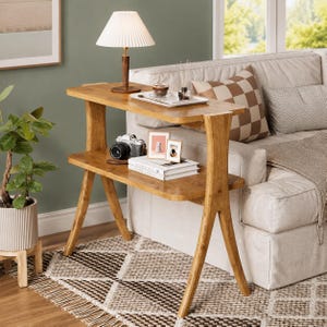 May include: A two-tier wooden console table with a light brown finish and angled legs. The top surface holds a pleated lamp, a tray with a glass, and a diffuser. The lower shelf displays a camera, two small framed pictures, and a stack of books.