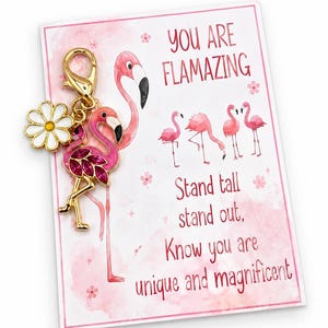 May include: A gold-tone metal flamingo charm with pink enamel and crystals, attached to a white daisy charm. It is displayed on a rectangular card featuring pink flamingo illustrations and the text: YOU ARE FLAMAZING, Stand tall, stand out, Know you are unique and magnificent.