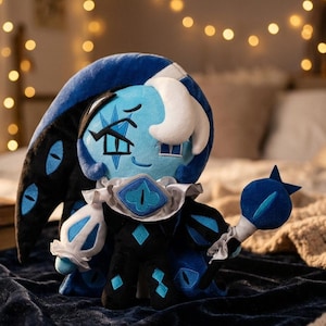 May include: A plush doll featuring a blue character with a white headpiece and a black and blue outfit. The doll has eye-shaped designs and is holding a blue staff with an eye-shaped design. The background is blurred with fairy lights.