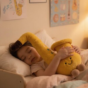 May include: A child asleep in bed, cuddling a large, yellow plush saxophone toy. The toy has a brown mouthpiece and green accents. The child is nestled under a blanket, with musical art on the wall.