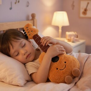 May include: A child asleep in bed, cuddling a plush orange ukulele toy. The toy has a smiling face and brown details. The scene is lit by a bedside lamp, with musical notes and framed art on the wall.