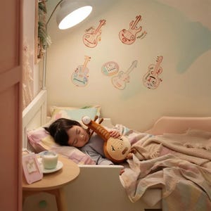 May include: A child sleeping in a bed, clutching a plush, brown, musical instrument toy. The wall is decorated with colorful guitar stickers. A small table holds a glass of milk and a picture frame.
