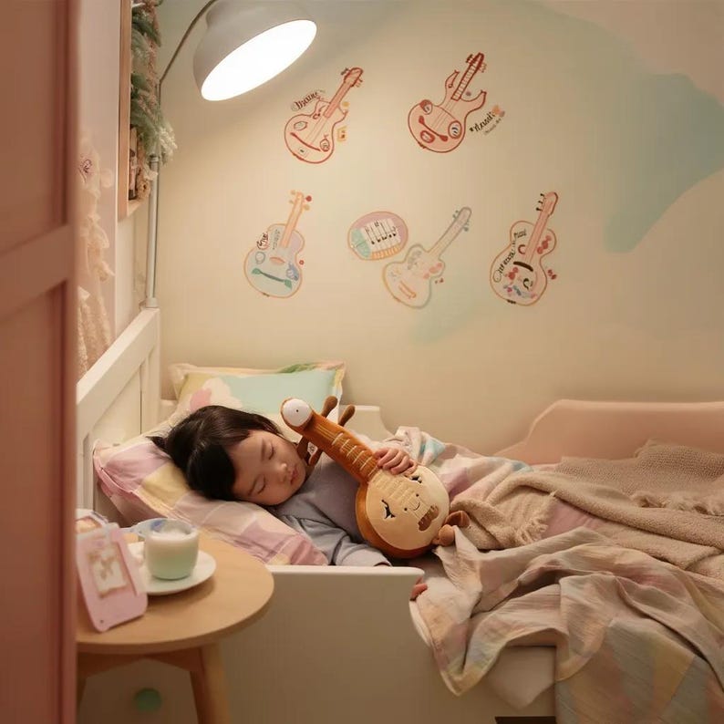 May include: A child sleeping in a bed, clutching a plush, brown, musical instrument toy. The wall is decorated with colorful guitar stickers. A small table holds a glass of milk and a picture frame.
