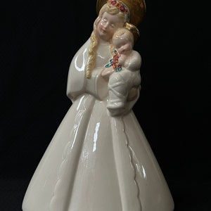 Pode incluir: Uma figurinha de cerâmica branca da Virgem Maria segurando um bebê Jesus. A figurinha está vestindo um vestido branco com uma auréola dourada e uma coroa de flores. O bebê Jesus está vestindo uma túnica branca e tem uma auréola dourada.