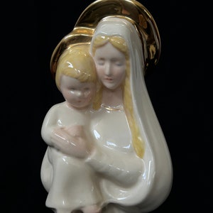 Pode incluir: Uma figurinha de cerâmica branca da Virgem Maria segurando um bebê Jesus. A figurinha é decorada com detalhes dourados e tem uma auréola ao redor da cabeça de Maria.