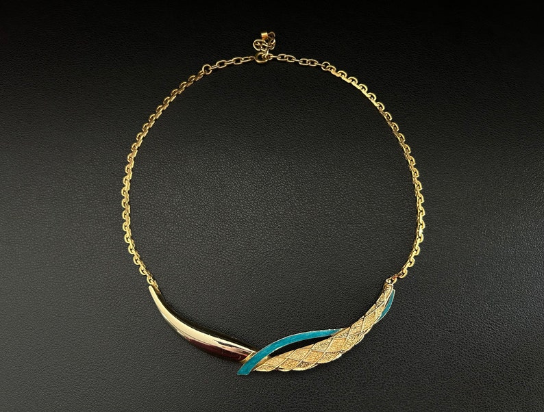 May include: A gold chain necklace with a blue and gold pendant. The pendant is shaped like a curved leaf with a woven design.