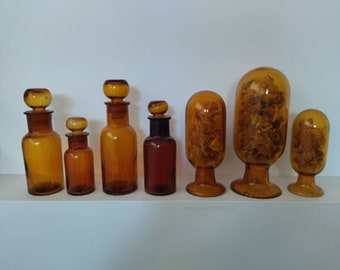 May include: A collection of seven amber glass bottles with various shapes and sizes. Some bottles have stoppers, while others have open tops. The bottles are arranged in a row on a white shelf.