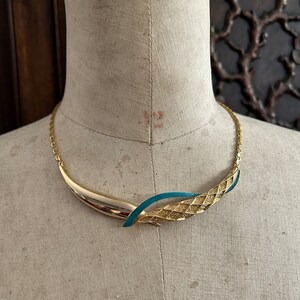 May include: A gold and teal blue statement necklace with a textured design. The necklace is on a mannequin.