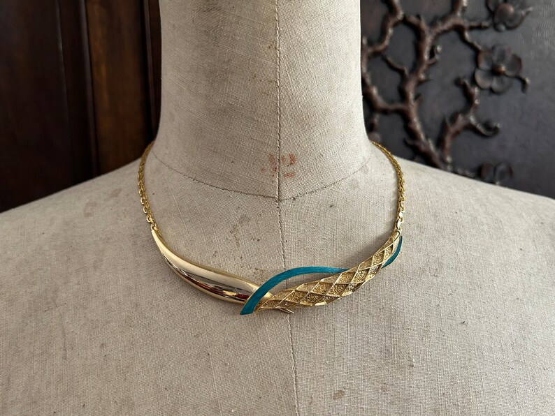 May include: A gold and teal blue statement necklace with a textured design. The necklace is on a mannequin.
