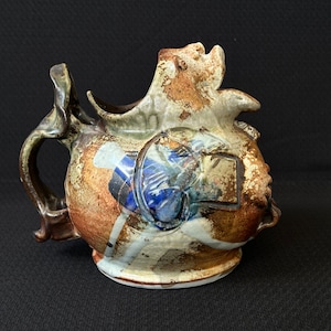 May include: A ceramic pitcher with a unique, abstract design. The pitcher is brown, white, and blue with a textured surface. It has a handle and a spout shaped like a face.